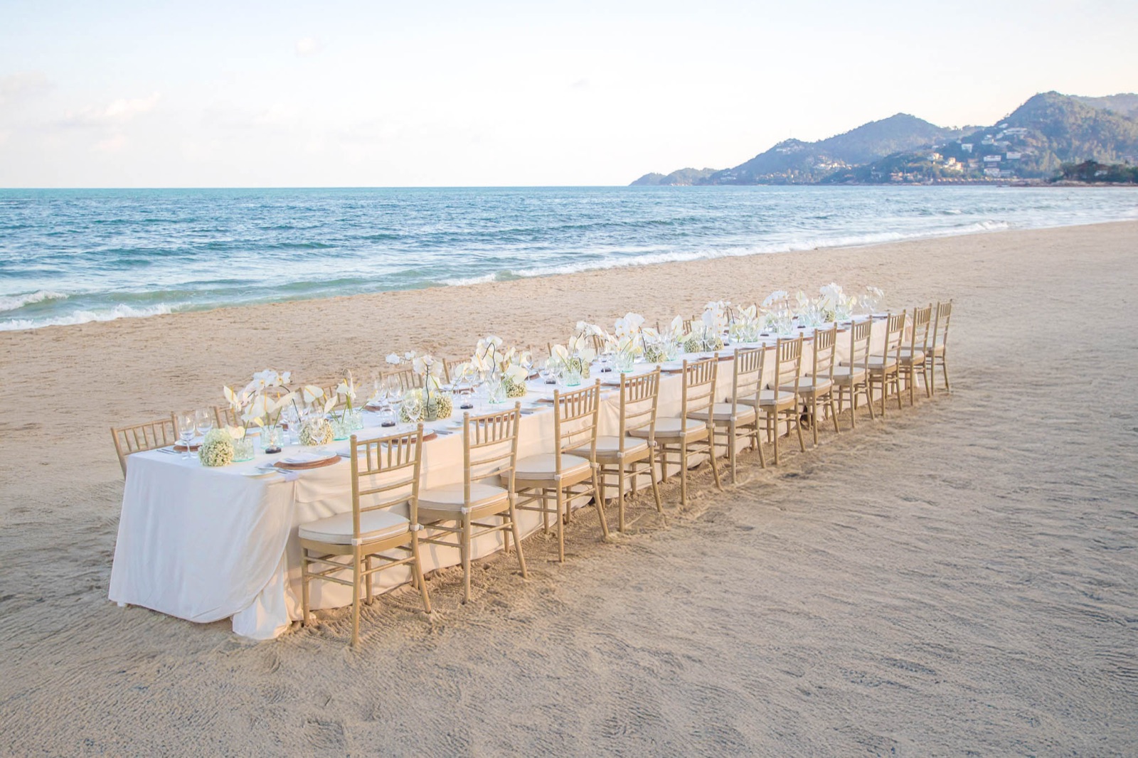 A long white dinner table set on the sand at Centara Reserve Samui, gold chairs and orchids, sea and headland at golden hour