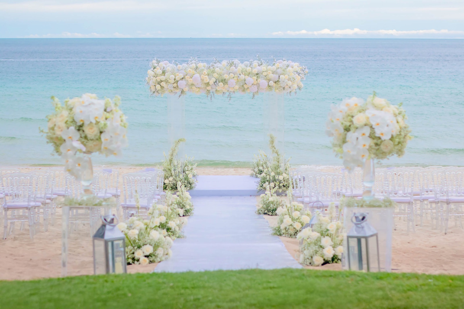 A white-rose ceremony arch on the garden lawn above the beach at Centara Reserve Samui, with clear chairs and lanterns