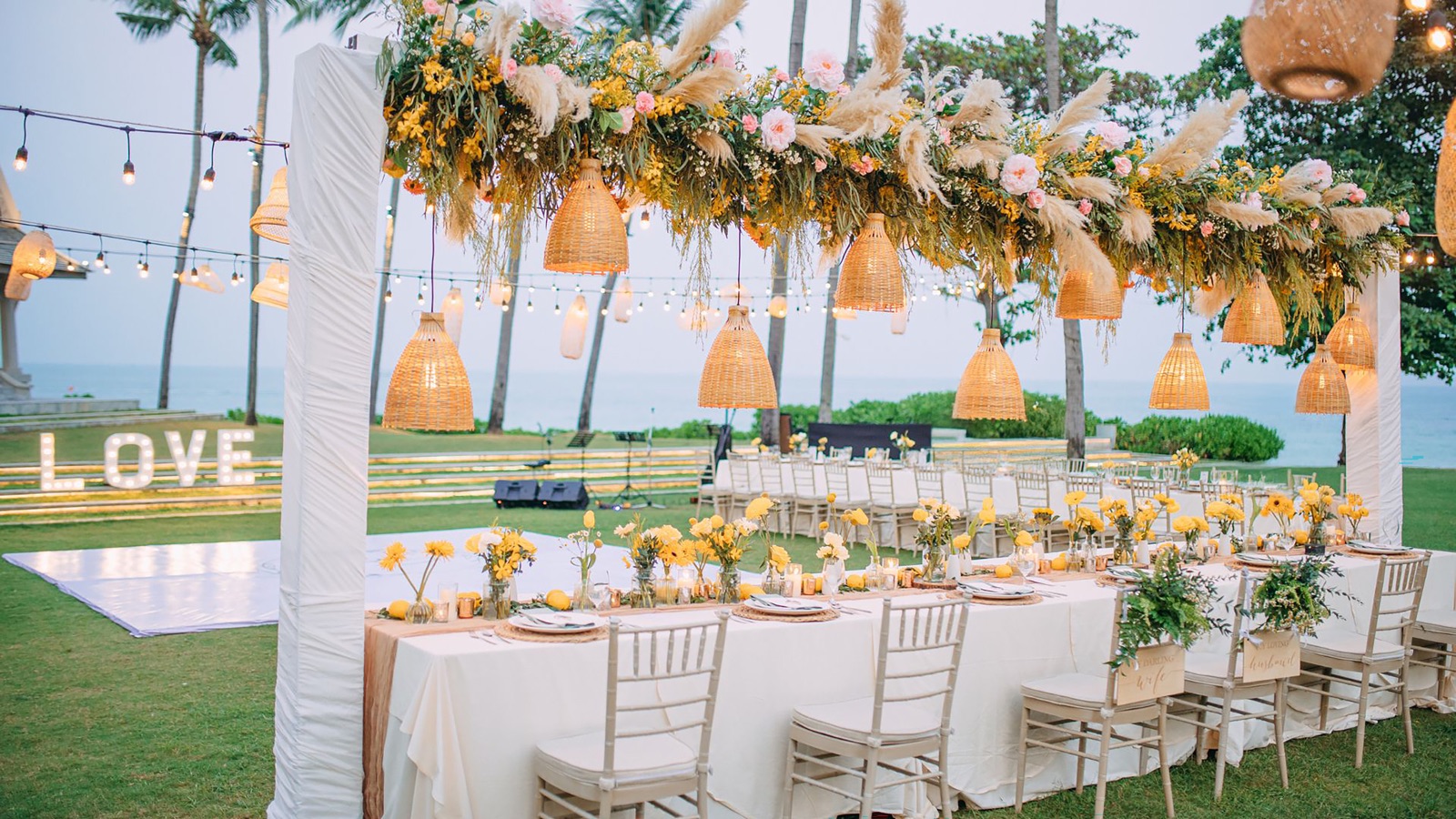 An evening wedding reception on the lawn at Centara Reserve Samui, with a pampas and floral canopy, rattan lanterns and a lit LOVE sign