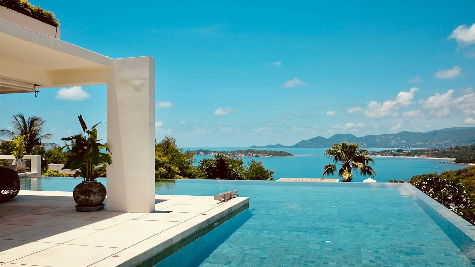A hillside villa terrace and infinity pool looking out over a blue bay and distant green hills