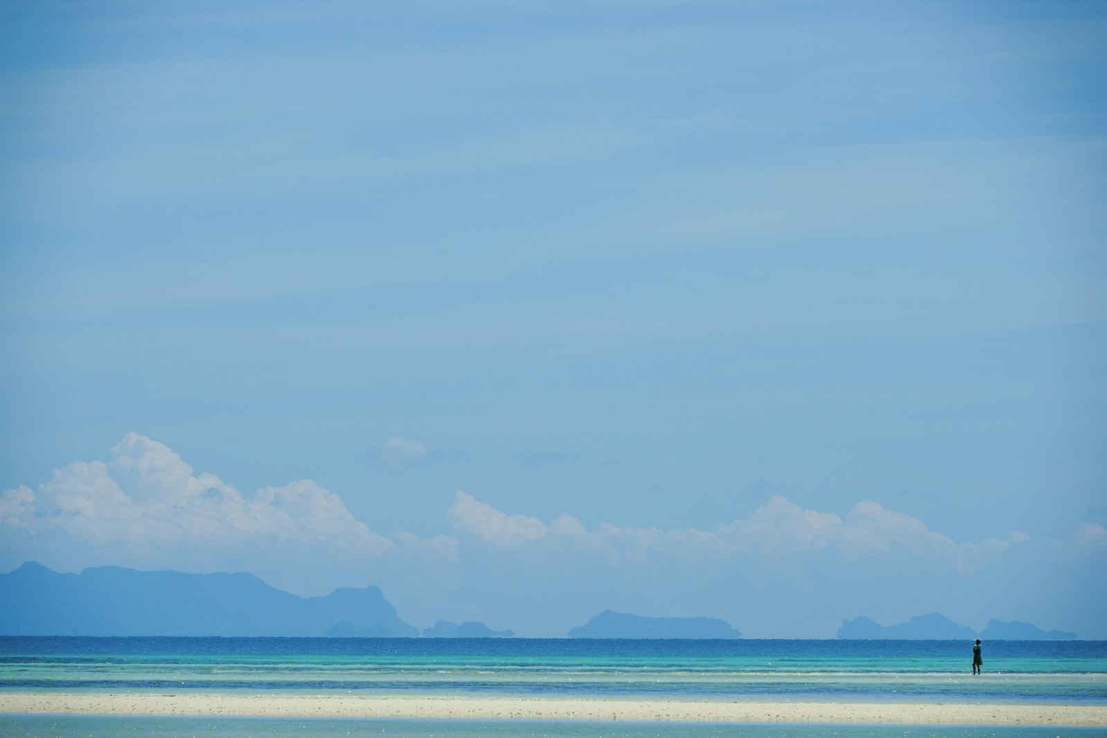 A single figure standing on an empty sandbar off Koh Samui, with a flat sea and distant islands on the horizon