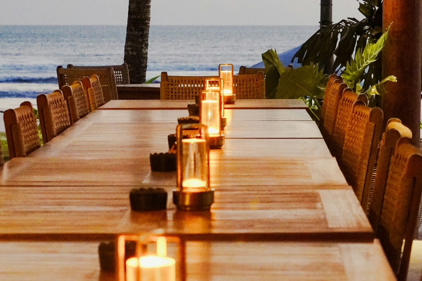 A long candle-lit table set with wicker chairs beside the sea at dusk, laid for a private dinner